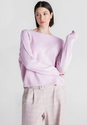 Vrouw met kort donker haar die een losse lichtroze trui en beige geruite broek draagt tegen een effen lichte achtergrond.