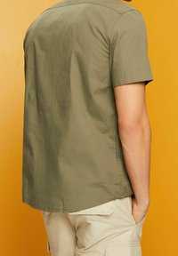 Man met een olijfgroen kortemouwenshirt en beige cargoshorts, staand met één hand in de zak tegen een gele achtergrond.