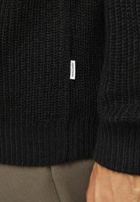 Maglione nero lavorato a maglia con texture a costine, dotato di un piccolo etichetta bianca. I polsini sono spessi e il tessuto appare caldo e accogliente.
