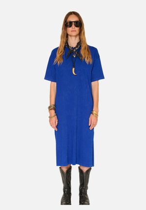 Femme portant une robe midi bleue, un bandana noir avec un collier, des lunettes de soleil noires, plusieurs bracelets et des bottes de cowboy noires, debout devant un fond blanc.