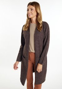 Cardigan lung de culoare maro, dintr-un material tricotat texturat, cu buzunare frontale, design deschis, purtat peste un top bej și pantaloni maro.