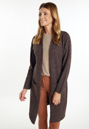 Brauner längerer Cardigan, strukturierter Strickstoff, mit aufgesetzten Taschen, offenes Design, getragen über einem beigen Oberteil und braunen Hosen.