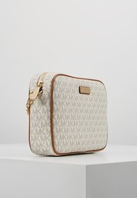 Kompaktowa beżowa torebka na ramię z brązowymi krawędziami, złotym zamkiem błyskawicznym oraz wzorem i emblematem MK, leżąca na białej powierzchni na jednolitym tle.