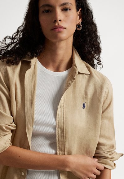 Kobieta z kręconymi włosami, ubrana w beżową koszulę na guziki na białej prążkowanej bluzce, podwijająca jeden rękaw, neutralne tło.