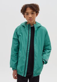 Teal windbreaker jas met capuchon, zwarte rits, elastische manchetten en zoom. Gelaagd over een zwart shirt; glad, lichtgewicht materiaal.