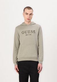 Olivgrüner Hoodie mit einer Vordertasche und einer Kapuze mit Kordelzug. Verziert mit dem "GUESS EST.1981"-Logo in dunklem Text. Lange Ärmel und strukturierten Stoff.