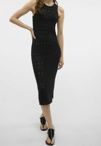 Černé pletené midi šaty s kulatým výstřihem, zdobené texturovaným vzorem a bočním rozparkem. Černé ploché sandály dotvářejí look.