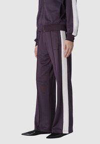 Paarse trainingsuit met witte zijstrepen, gemaakt van een gladde stof. De broek heeft een losse pasvorm en wordt gecombineerd met puntige zwarte schoenen.