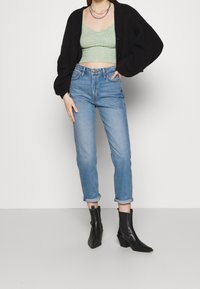 Persona que lleva un cárdigan negro, una camiseta verde cortada, unos vaqueros azules de tiro alto y remangados, y botas de tobillo negras de punta.}}