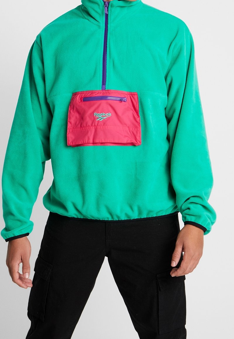 Grüner Fleece-Pullover mit halbem Reißverschluss, ausgestattet mit einer pinken Fronttasche und Logo. Elastische Bündchen und Saum, weiche Textur und lässige Passform.