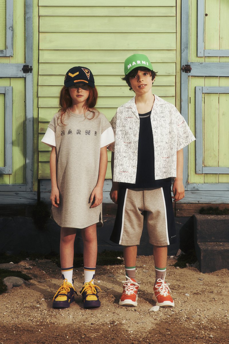 Två barn står framför en grön trälängd. Flickan har på sig en grå klänning med vita ärmar och pojken har en mönstrad t-shirt med korta ärmar. Båda har färgglada sneakers.