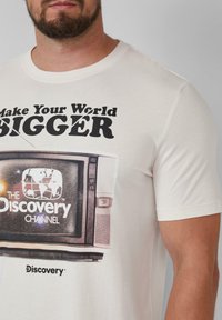 Witte katoenen t-shirt met een graphic van een vintage televisie met de tekst "Maak jouw wereld GROTER" en het logo van "The Discovery Channel".