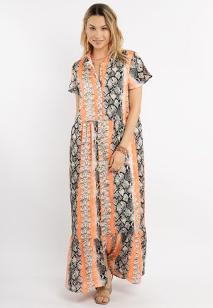 Vrouw draagt een lange jurk met korte mouwen met verticale oranje, zwarte en witte slangenhuidprint strepen, staand en glimlachend.