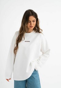 Witte sweatshirt met lange mouwen en een ronde halslijn, met geborduurd "MIRONS" in bruin. Gecombineerd met blauwe wijde jeanstroef. Zachte textuur.