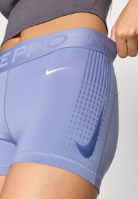 Nike Pro šortai šviesiai purpurinės spalvos su elastingu juosmeniu, dekoruoti iškilniu logotipu ir horizontaliais juostelėmis. Lygus, elastingas audinys.
