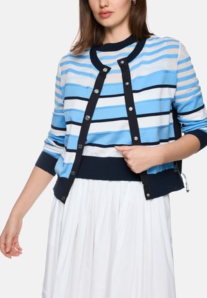 Woman wearing a blue, white, and black striped buttoned cardigan over a matching striped top and a white pleated skirt on a plain background.