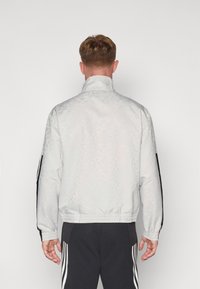 Giacca bomber bianca con una texture a motivi sottili, strisce laterali nere, polsini elastici e cintura elasticizzata; vista da dietro.