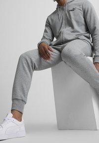 Sweat à capuche gris zippé et pantalon de survêtement assorti en tissu doux, avec poignets côtelés et petit logo noir sur la poitrine. Baskets blanches.