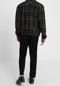 Giacca a quadri nera e grigia con colletto classico, indossata sopra pantaloni neri affusolati, abbinata a scarpe nere. Tessuto texture, design casual.