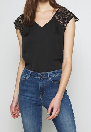 Kvinde iført en sort ærmeløs top med blonder på skuldrene sammen med blå, højttaljede jeans, stående mod en ensfarvet baggrund.