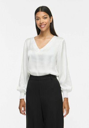 Jeune femme aux longs cheveux foncés souriante, portant un chemisier blanc à col en V avec des manches longues bouffantes et un pantalon noir taille haute.