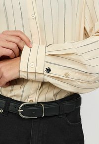 Tejfehér ing függőleges fekete csíkokkal, gombos mandzsettával, és egy kis hímzett logóval az ujjon. Fekete texturált öv látható.