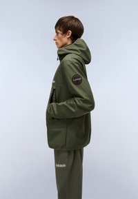 Olivengrøn jakke med hætte, med lynlåslommer og en sort cirkulær logo-patch på ærmet; parret med matchende bukser.