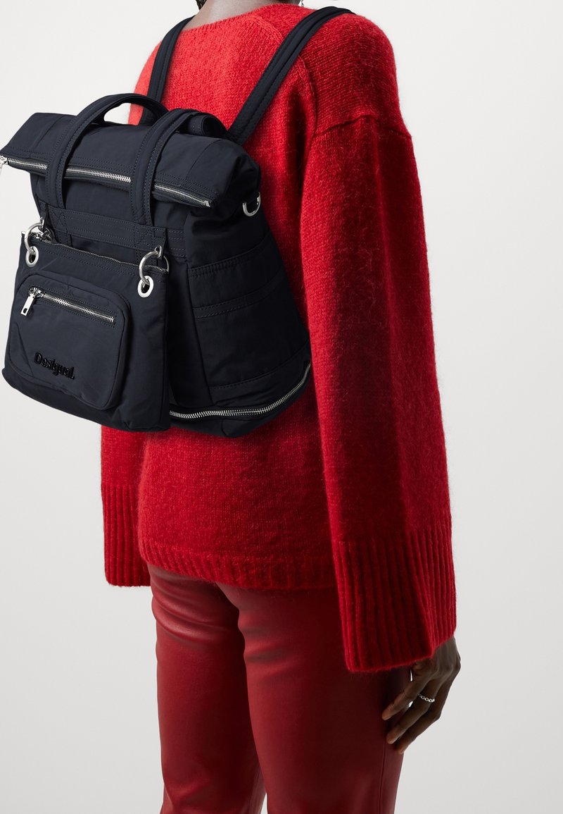 Personne portant un pull rouge et un pantalon, portant un sac à dos noir avec plusieurs fermetures éclair dans le dos sur un fond uni.
