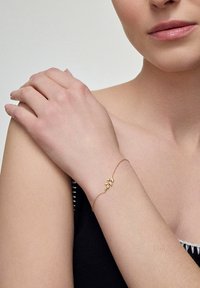 Złota bransoletka łańcuszkowa z delikatnym wzorem liścia, noszona na jasnej skórze nadgarstka na neutralnym tle. Gładka tekstura, minimalistyczny styl.