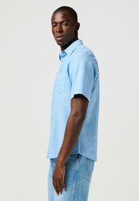 Camicia azzurra a maniche corte con colletto button-down, tessuto strutturato, taschino sul petto e orlo curvo, abbinata a jeans in denim chiaro.