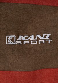 Brun og rødstripet stoff med myk tekstur. Fremtredende hvit "KANI SPORT" logo brodert på den brune delen.