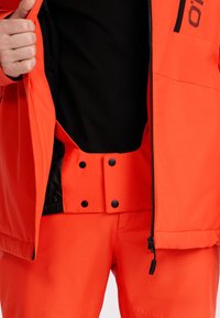 Veste extérieure orange vif avec des accents noirs, dotée d'une fermeture éclair, de boutons-pression et d'un intérieur en mesh. Associée à un pantalon assorti.