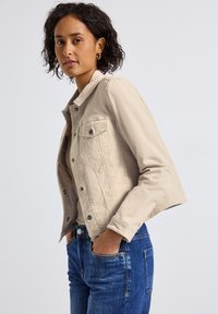 Veste en denim beige avec col, boutons sur le devant et deux poches poitrine. Associée à un jean bleu, avec une coupe droite et des poches latérales.