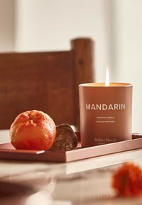 Mandarinen-Duftkerze in einem matten pfirsichfarbenen Behälter mit weißem Text, begleitet von einer Orange und einem dunklen Stein, auf einem rosa Tablett.