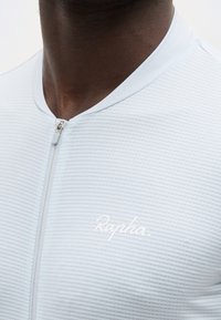 Κοντινό πλάνο ενός ατόμου που φοράει ανοιχτό γκρι ποδηλατική μπλούζα με φερμουάρ και διακριτικές οριζόντιες ρίγες, με το λογότυπο "Rapha" στο στήθος.