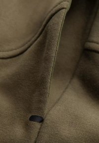 Tissu de sweatshirt vert olive avec une texture douce, présentant une couture proéminente et un petit détail noir près du bord.