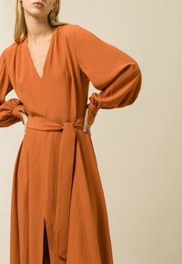 Dlouhé oranžové šaty s výstřihem do V, nafouknutými rukávy a zavázaným pasem. Mají rozparek vpředu a hladkou, splývavou texturu látky.