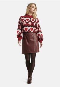 Red and pink heart-patterned knitted sweater with puff sleeves paired with a burgundy faux leather mini skirt and black tights. Glossy ankle boots.