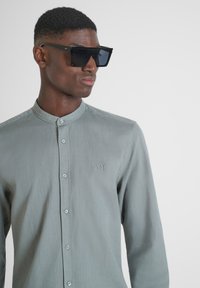 Camicia grigia con colletto alla mandarino e una sottile trama, dotata di un logo ricamato e bottoni azzurri chiari. Grandi occhiali da sole neri.