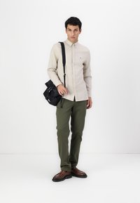 Cămașă bej deschis cu nasturi, pantaloni verzi măslinii, geantă de umăr neagră și pantofi maro. Design curat, cu accente și texturi minime.