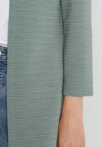 Ärmen på en ribbad, salviagrön kofta som bärs över en vit skjorta och blå jeans, med detaljer av armen och sidotrycket synliga.