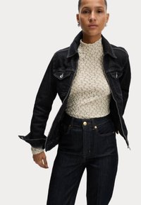 Svart denimjacka med två bröstfickor och silverdetaljer, lager på lager över en ljus, mönstrad polotröja och högmidjade mörka jeans.