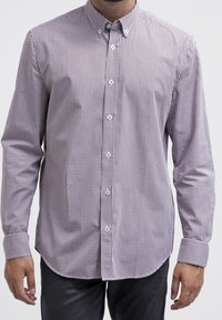 Mann in einem langen, knopfgebundenen, kleinen lila-weiß karierten Hemd mit Kragen und Manschetten, kombiniert mit dunklen Hosen.