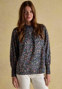 Jauna sieviete ar garām brūnām matiem, valkājot tumšu ziedu blūzi ar smocka detaļām un baltām biksēm, stāv uz beigē fona.