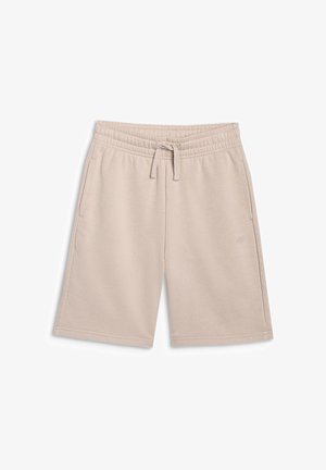 Garçon portant un t-shirt noir à manches longues, un short beige, des chaussettes blanches et des baskets grises, debout les mains dans les poches.