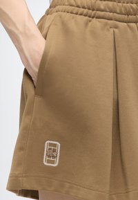 Gros plan d'une main dans la poche d'un short marron texturé avec une ceinture élastique et un petit logo brodé près de l'ourlet.