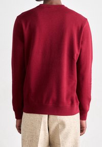 Sweat-shirt bordeaux à manches longues, col rond et ourlet côtelé. Texture lisse, silhouette ajustée, porté avec un pantalon beige texturé.