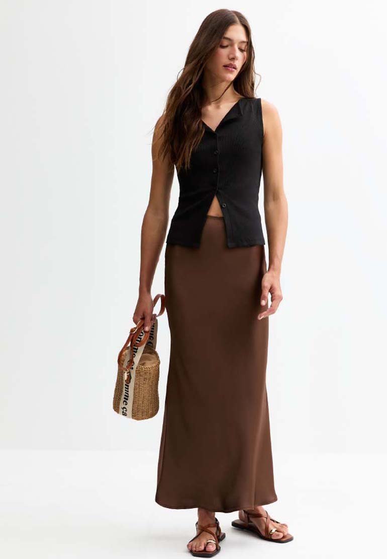 Haut noir sans manches avec des boutons, jupe brune à la cheville et sandales plates brunes. Le modèle tient un sac en osier avec une bandoulière.