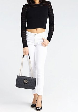 Femme portant un crop top noir transparent à manches longues, un jean skinny blanc, des talons noirs ornés et tenant un sac à main noir avec des anses en chaîne dorée.