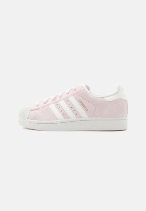 Jasnoróżowy but sportowy Adidas Superstar z białymi paskami, białymi sznurowadłami i nakładką na palce, pokazany z boku na jednolitym tle.